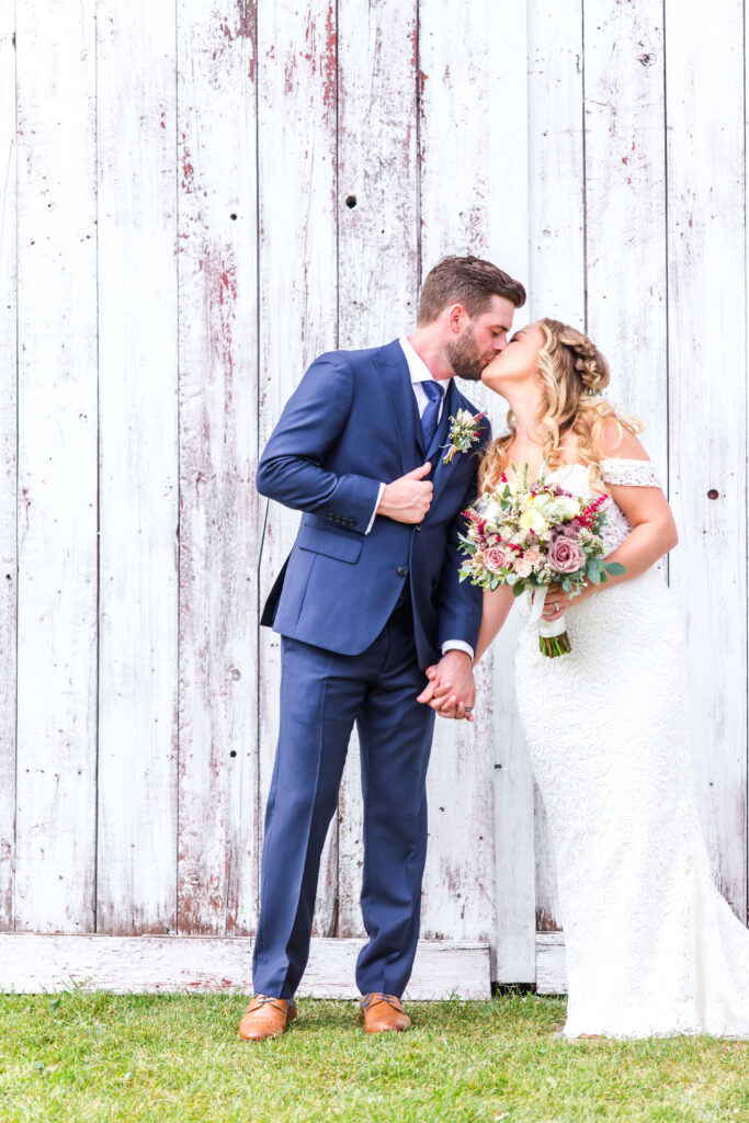 Bride & Groom share a kiss in front of white barn at summer wedding at farmin' betty's 